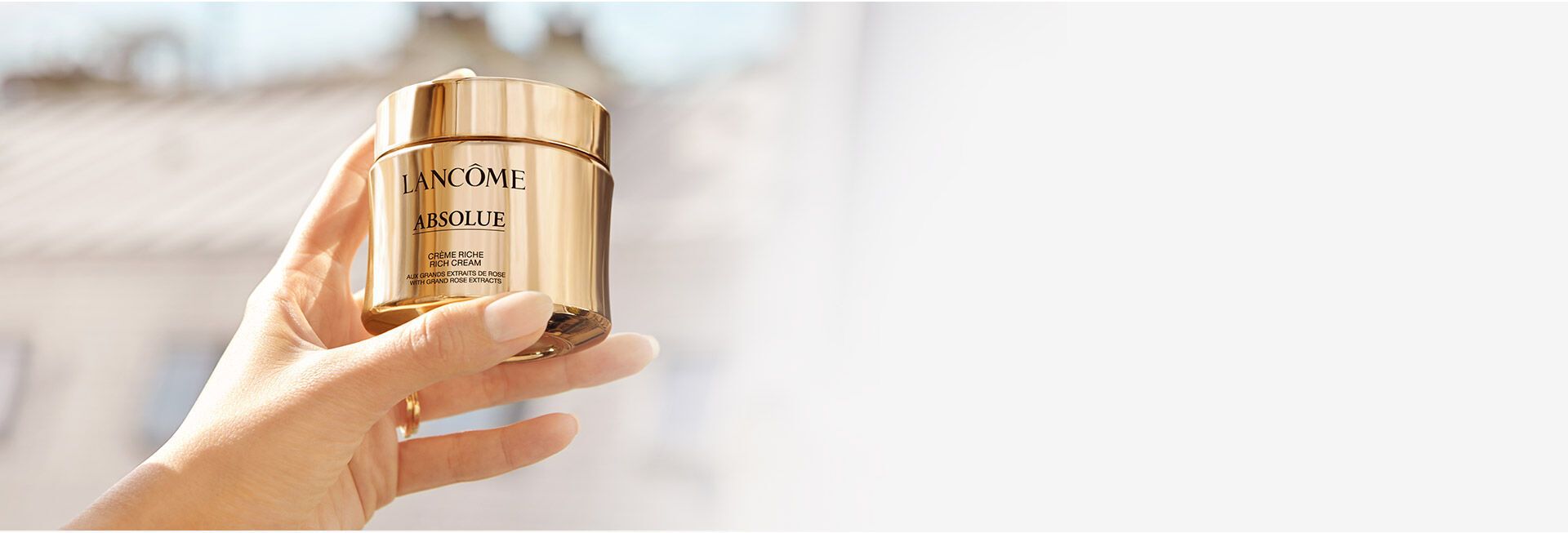 A hand holding a luxurious gold jar of Lancome Absolue skincare cream, with a soft, blurred background suggesting natural light.
