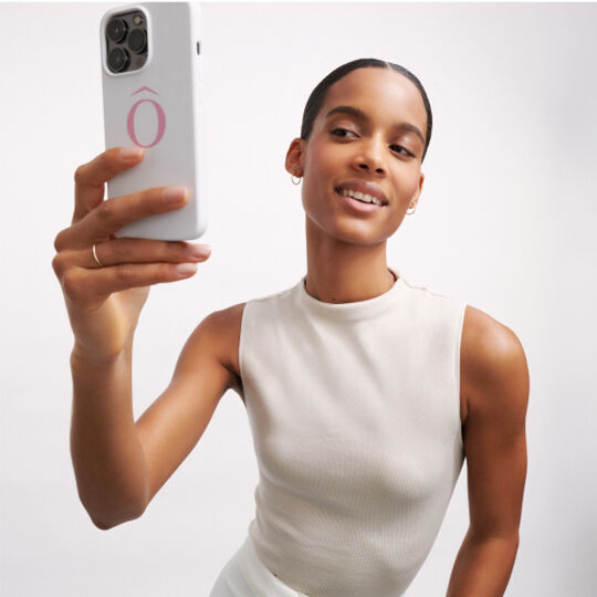 Een vrouw maakt een selfie met een smartphone, terwijl ze glimlacht naar de camera. Ze draagt een lichtgekleurde top en heeft een strakke haarstijl.