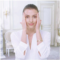 A woman in a white blouse is applying gentle pressure to her face with her hands, smiling softly. She is in a bright, elegantly decorated room.