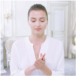 A woman with her eyes closed, wearing a white top, is gently clasping her hands together in a serene setting.