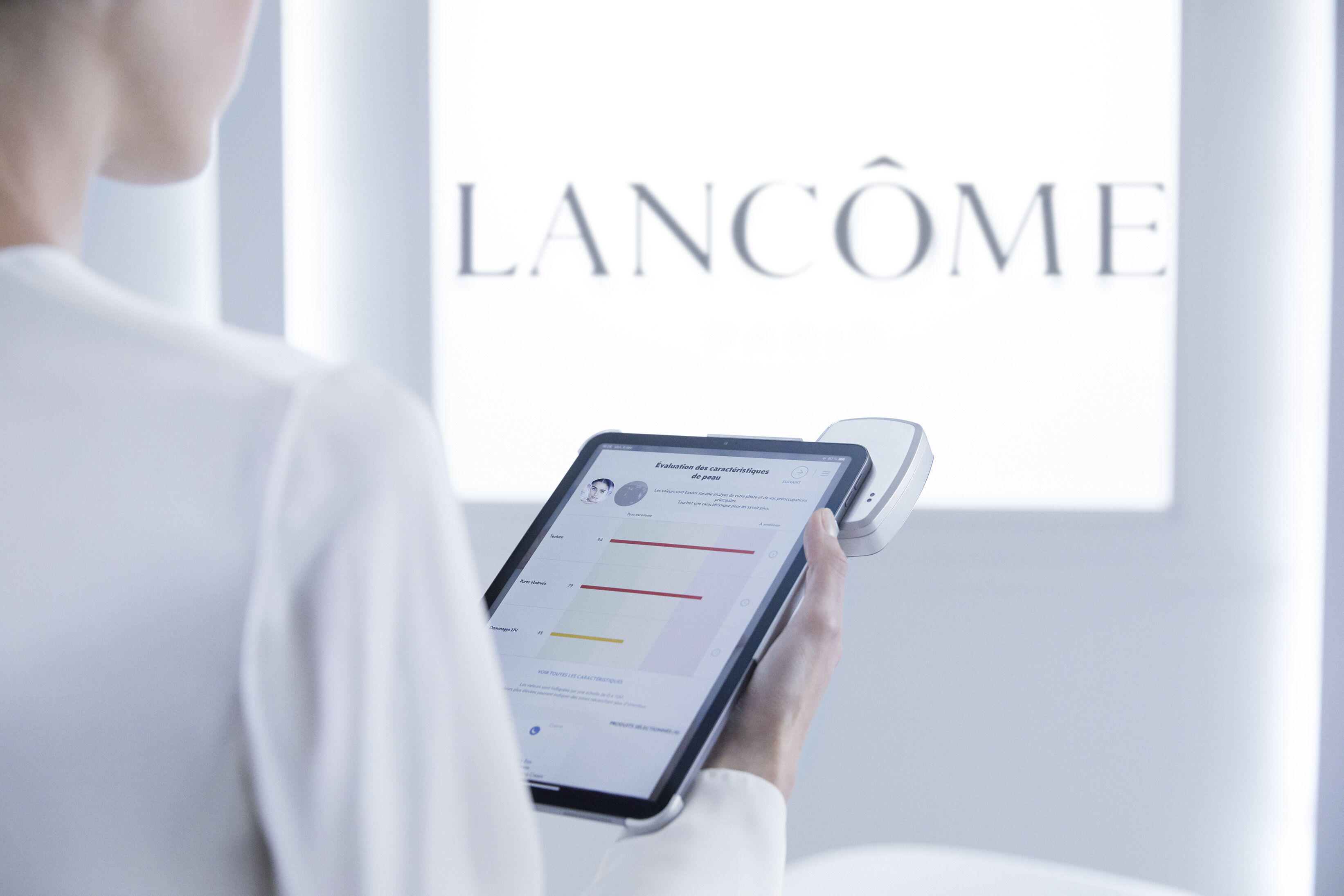 Een persoon houdt een tablet in de hand met huidanalysegegevens, terwijl op de achtergrond het Lancome-logo zichtbaar is.