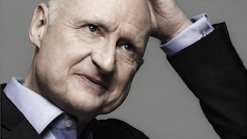 A man in a suit is slightly tilting his head and touching his forehead with his hand, looking thoughtfully at the camera.