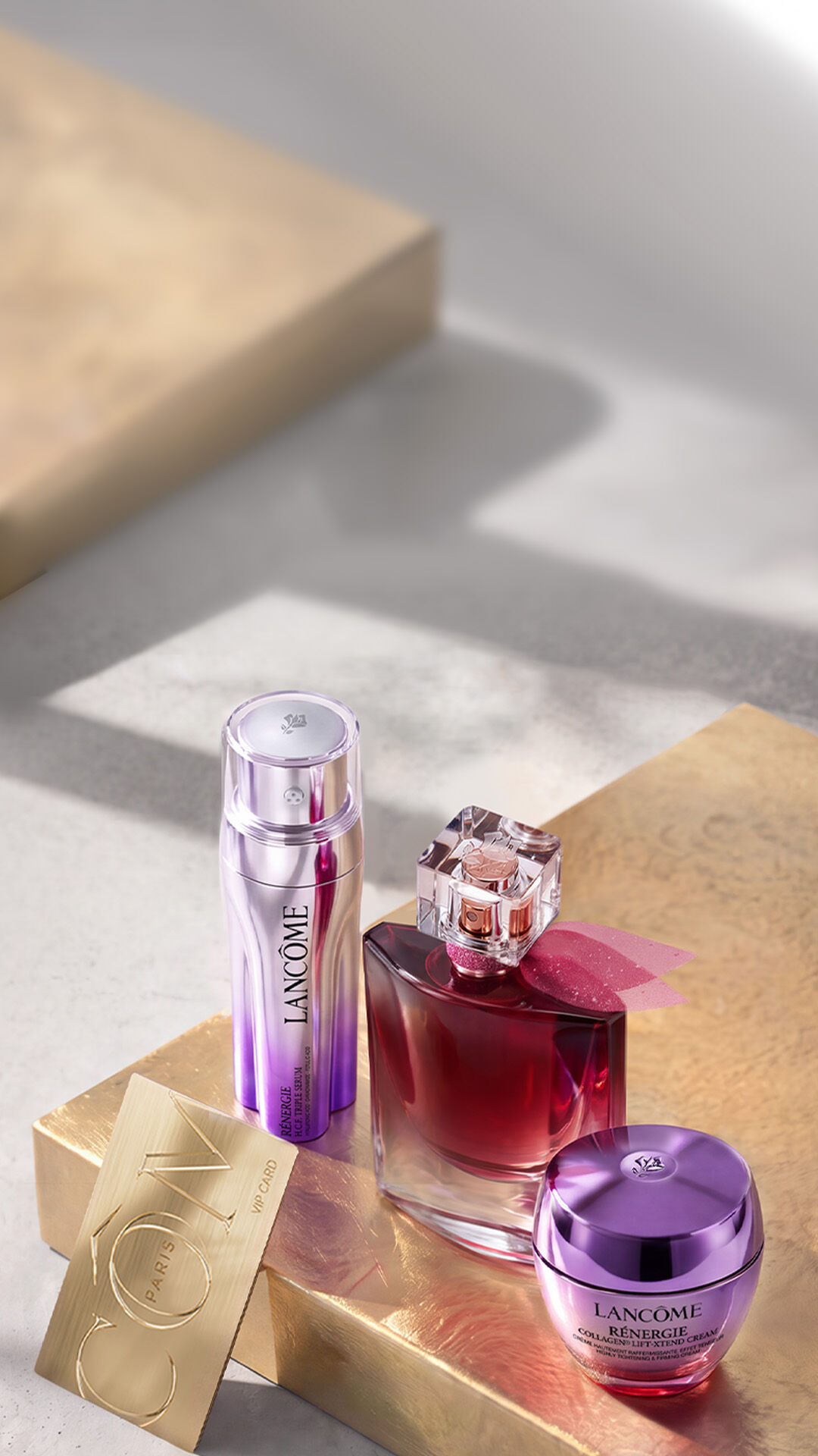 Renergie skincareproducten, waaronder een oogcreme, anti-aging creme en serum, gepresenteerd op een glanzend roze oppervlak voor een roze villa met cipressen en een blauwe hemel.