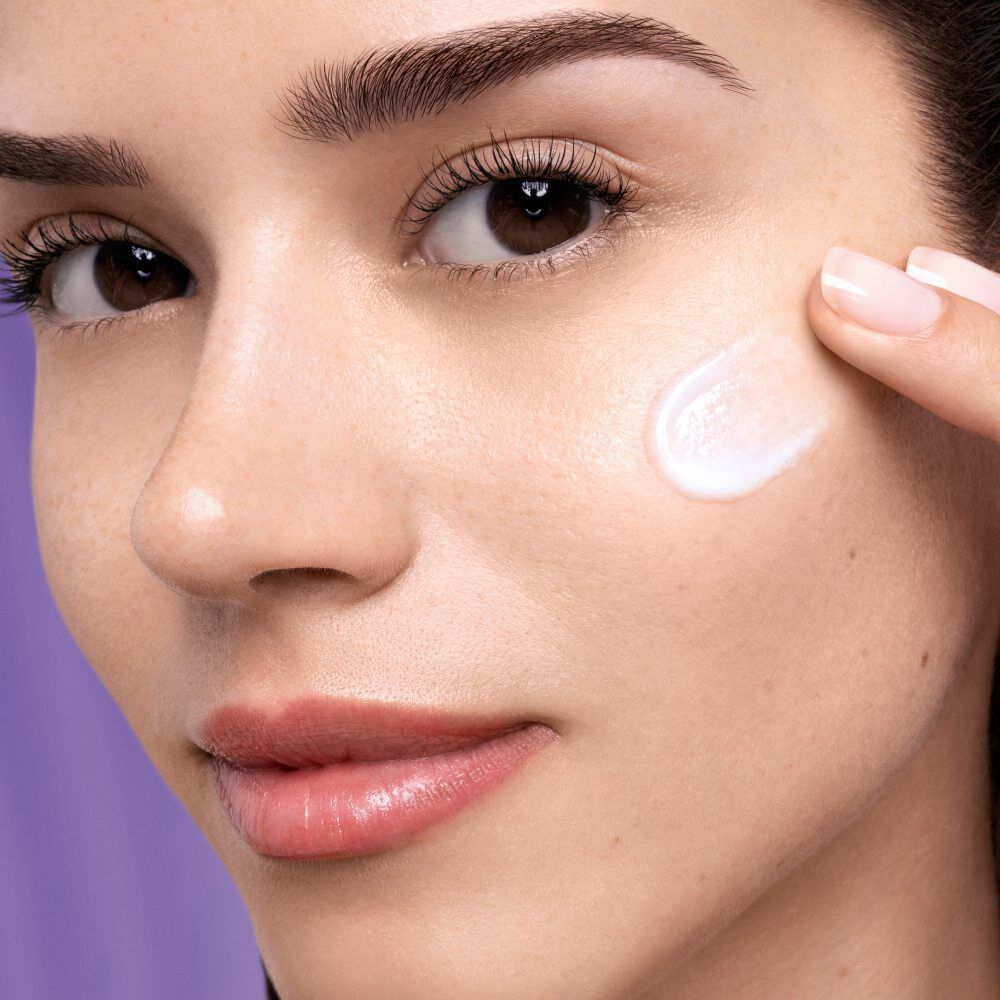 Een vrouw met een frisse huid brengt een witte creme aan op haar wangen tegen een paarse achtergrond.