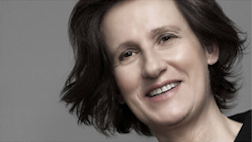 A smiling woman with medium-length wavy hair, wearing a black top, against a gray background.