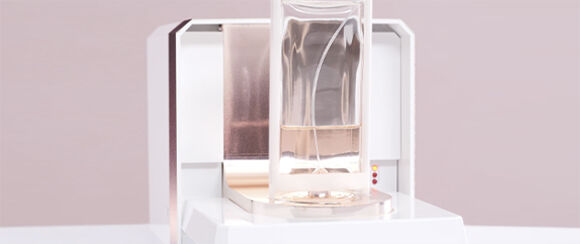 A sleek, modern device with a cylindrical glass container positioned on top, set against a soft pink background.