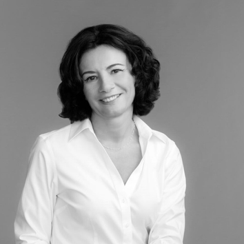 Zwart-wit portret van een vrouw met krullend haar, glimlachend in een witte blouse tegen een neutrale achtergrond.