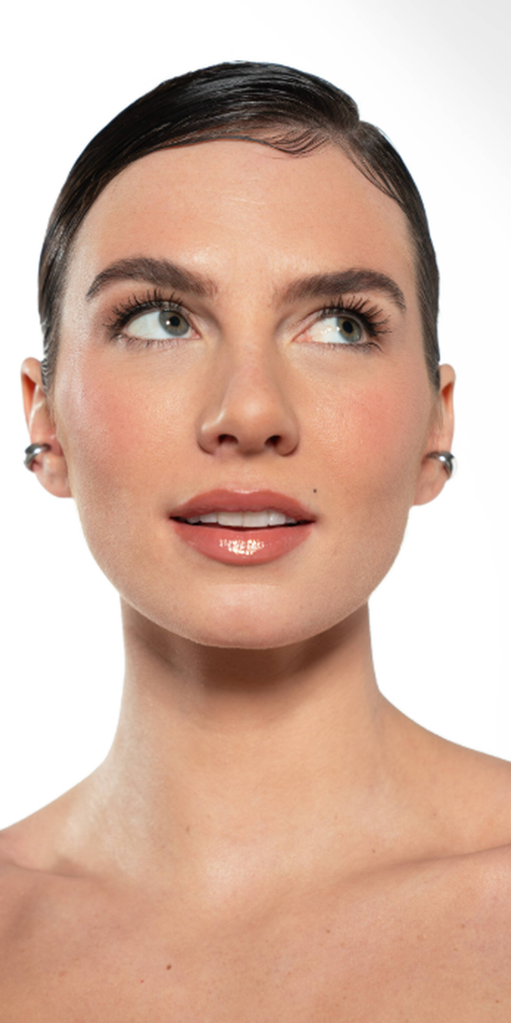Een vrouw met een gladde, gepolijste look, kort haar en een frisse make-up, kijkt omhoog met een dromerige uitdrukking. Ze draagt een glanzende zwarte top en heeft subtiele oorbellen.