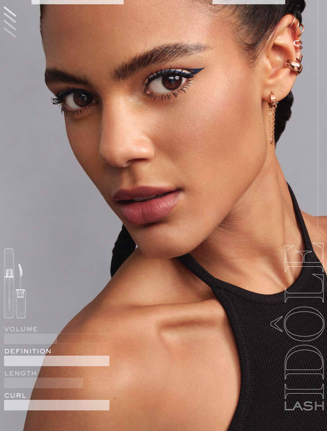 Een close-up van een vrouw met een strakke eyeliner en volle lippen, gekleed in een zwarte top. Haar haar is in een vlechtstijl, met een opvallende oorbel. De achtergrond is neutraal en er staan tekstlabels voor volume, definitie, lengte en krul in de hoek.