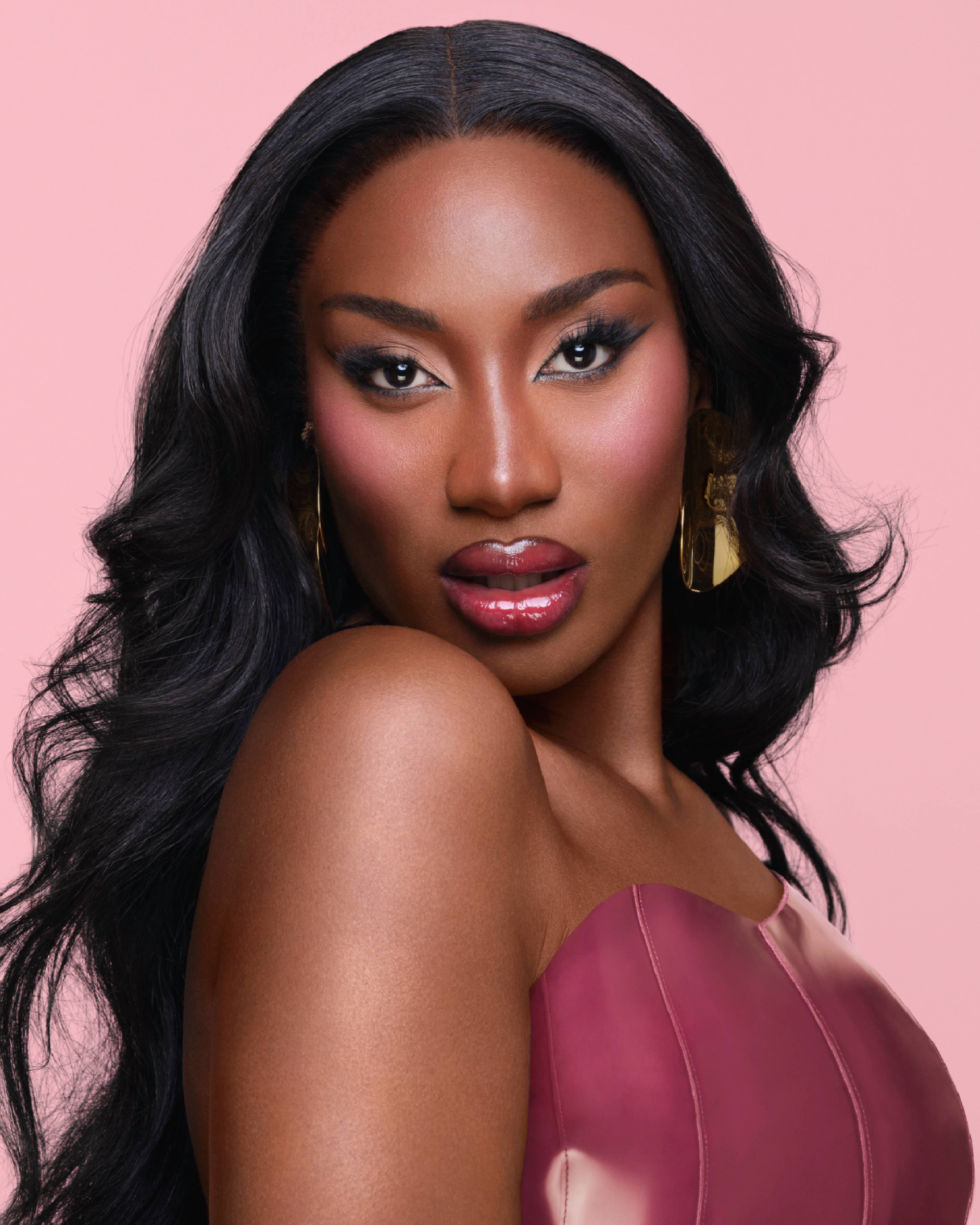 Een portret van een vrouw met een glanzende huid, lange donkere haren en opvallende make-up. Ze draagt een strapless top en grote oorbellen, met een roze achtergrond.